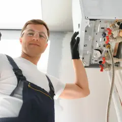 Installatore sorridente con occhiali protettivi e guanti fa segno OK vicino a una caldaia a compensazione appena installata.