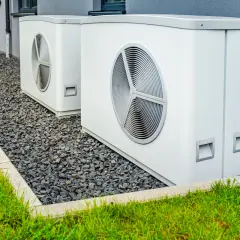 Due unità esterne di pompe di calore alta temperatura, bianche e moderne, installate su ghiaia accanto a un prato verde e a un edificio grigio.
