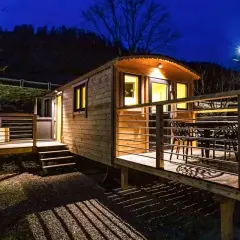 Caravan in legno leggero e resistente, illuminata di notte, con terrazza e tavolo esterno.