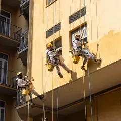 Lavoratori in corda applicano intonaco esterno tipi su un edificio giallo.