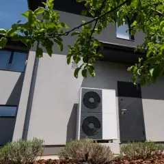 Unità esterna di una pompa di calore moderna, installata accanto a una casa con finiture grigie e vegetazione rigogliosa.
