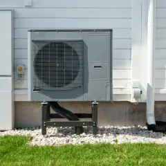 Unità esterna per riscaldamento ad aria, montata su supporti, accanto a un armadio tecnico bianco su una casa con rivestimento in legno.