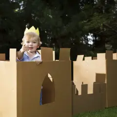 Bambino con corona gioca in un castello fatto con giochi fai da te con cartone, in giardino.