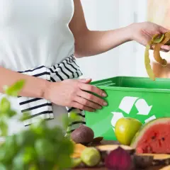 Mani che gettano bucce di frutta in un contenitore verde per ridurre i rifiuti. Frutta e verdura sul tavolo.