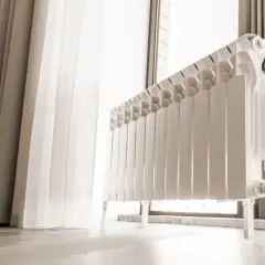 Radiatore bianco vicino a una finestra con tenda. Valutare il teleriscaldamento pro e contro per un riscaldamento efficiente.