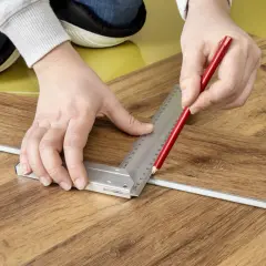 Mani che segnano con matita e squadra un listone per la posa pavimento pvc effetto legno.