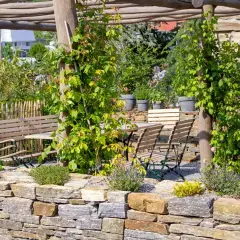 Un angolo di giardino con muretti a secco, un pergolato in legno, tavoli e sedie, e un'aiuola fiorita.