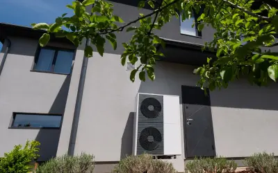 Unità esterna di una pompa di calore moderna, installata accanto a una casa con finiture grigie e vegetazione rigogliosa.