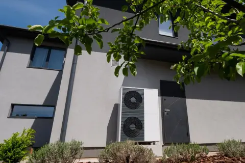 Unità esterna di una pompa di calore moderna, installata accanto a una casa con finiture grigie e vegetazione rigogliosa.