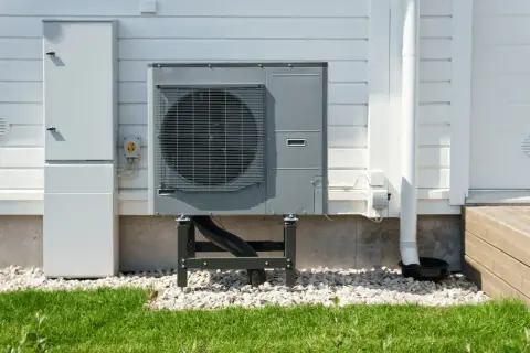 Unità esterna per riscaldamento ad aria, montata su supporti, accanto a un armadio tecnico bianco su una casa con rivestimento in legno.