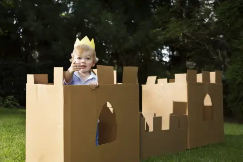 Bambino con corona gioca in un castello fatto con giochi fai da te con cartone, in giardino.