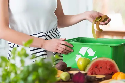 Mani che gettano bucce di frutta in un contenitore verde per ridurre i rifiuti. Frutta e verdura sul tavolo.