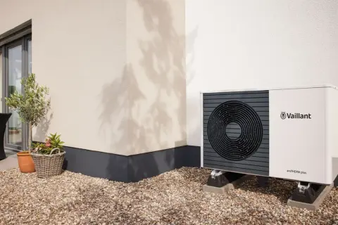 Pompa di calore climatizzatore Vaillant aroTHERM plus, un'unità esterna moderna su un patio ghiaioso, con un piccolo ulivo e un cesto di fiori.