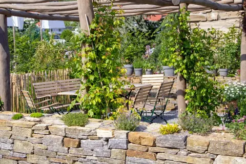 Un angolo di giardino con muretti a secco, un pergolato in legno, tavoli e sedie, e un'aiuola fiorita.