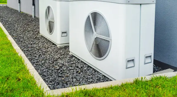 Due unità esterne di pompe di calore alta temperatura, bianche e moderne, installate su ghiaia accanto a un prato verde e a un edificio grigio.