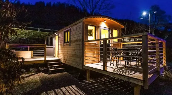 Caravan in legno leggero e resistente, illuminata di notte, con terrazza e tavolo esterno.