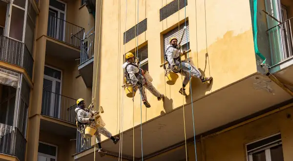 Lavoratori in corda applicano intonaco esterno tipi su un edificio giallo.