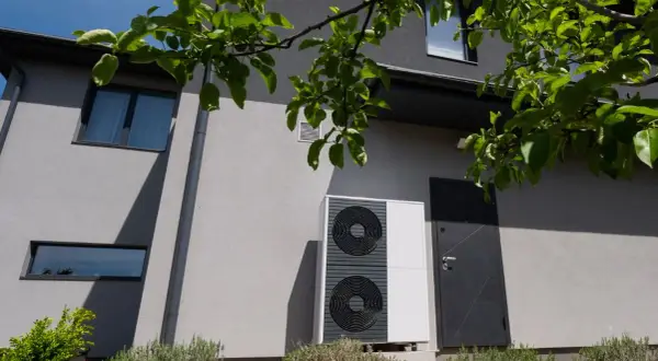 Unità esterna di una pompa di calore moderna, installata accanto a una casa con finiture grigie e vegetazione rigogliosa.