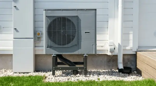 Unità esterna per riscaldamento ad aria, montata su supporti, accanto a un armadio tecnico bianco su una casa con rivestimento in legno.