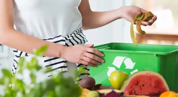 Mani che gettano bucce di frutta in un contenitore verde per ridurre i rifiuti. Frutta e verdura sul tavolo.