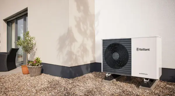 Pompa di calore climatizzatore Vaillant aroTHERM plus, un'unità esterna moderna su un patio ghiaioso, con un piccolo ulivo e un cesto di fiori.