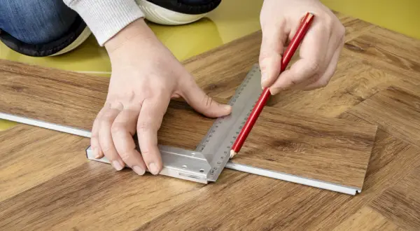 Mani che segnano con matita e squadra un listone per la posa pavimento pvc effetto legno.