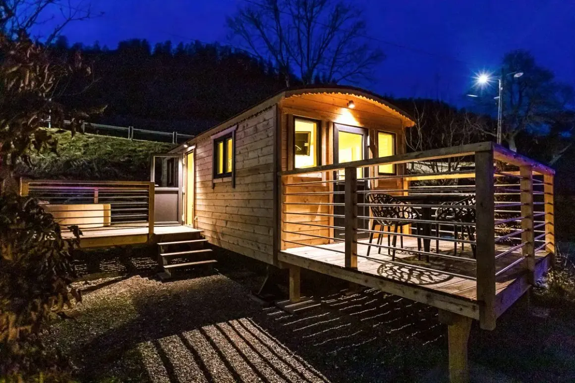 Caravan in legno leggero e resistente, illuminata di notte, con terrazza e tavolo esterno.