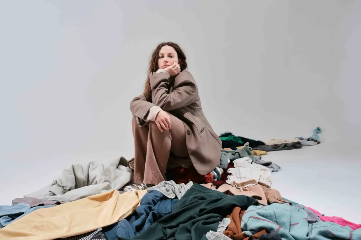Donna pensierosa seduta su un cumulo di vestiti, simbolo del consumismo del brand fast fashion.