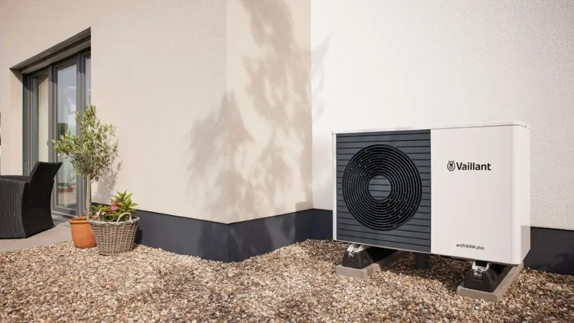 Pompa di calore climatizzatore Vaillant aroTHERM plus, un'unità esterna moderna su un patio ghiaioso, con un piccolo ulivo e un cesto di fiori.