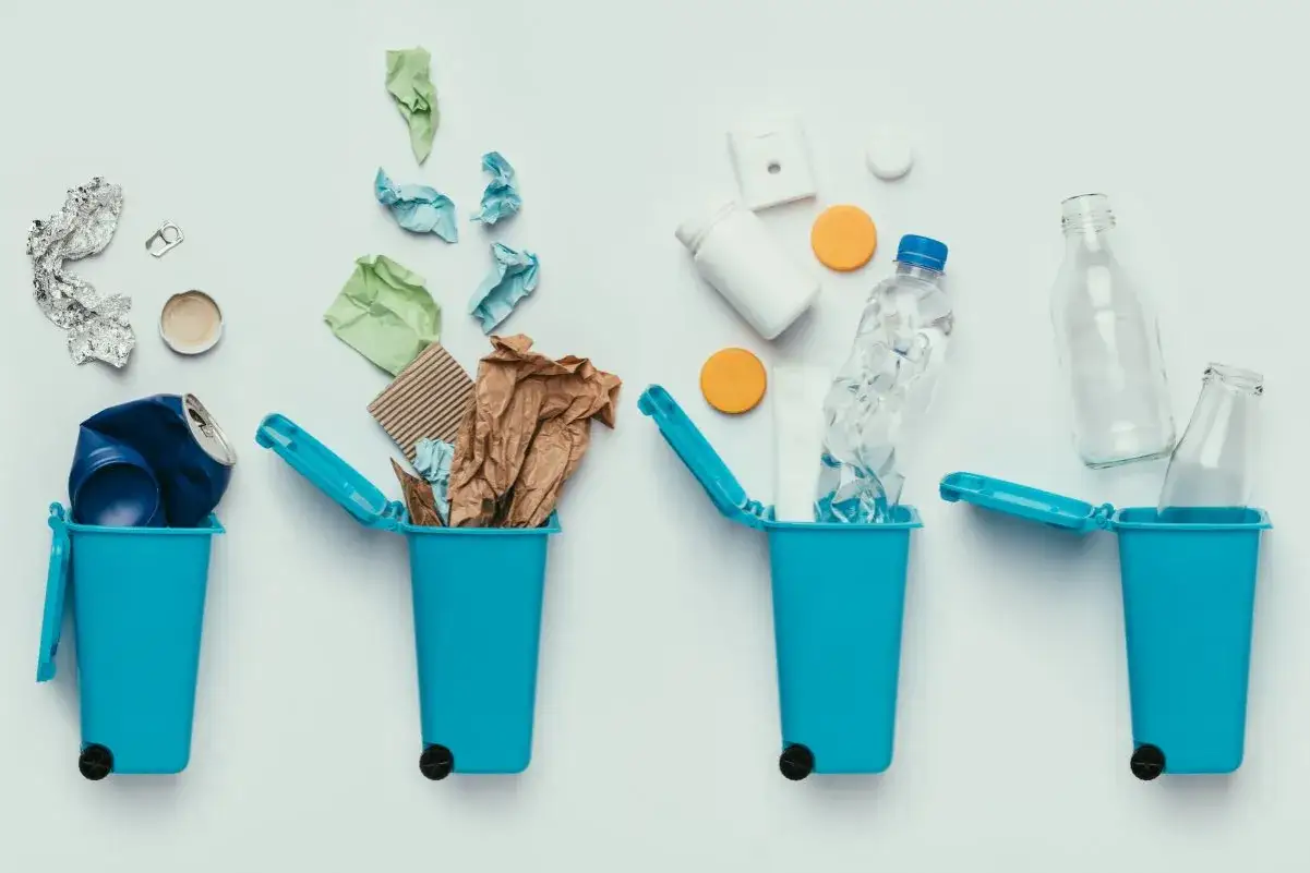 Quattro bidoni blu contengono rifiuti: lattine, carta, bottiglie di plastica e vetro. Oltre 20 oggetti di plastica e altri materiali pronti per il riciclo.