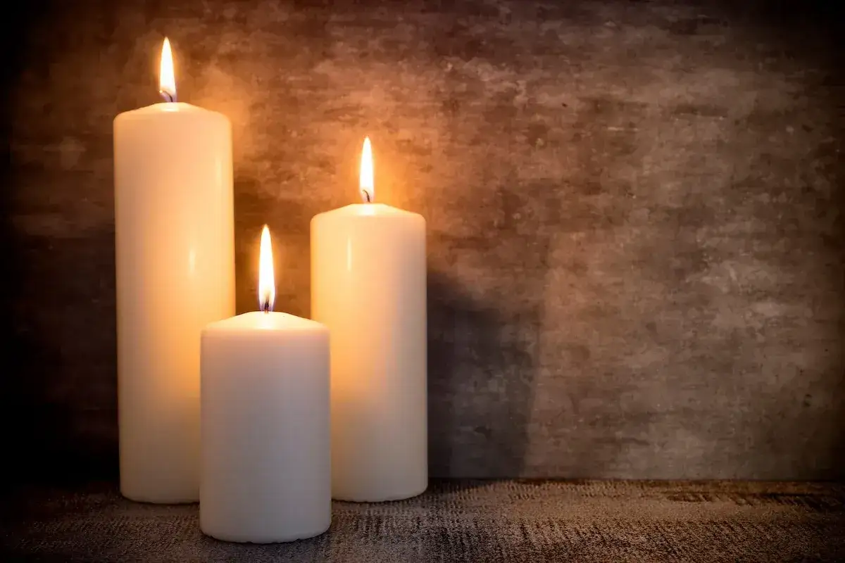 Tre candele di cera bianca brillano su uno sfondo scuro. Quando si consumano, ci si chiede dove si buttano le candele di cera.