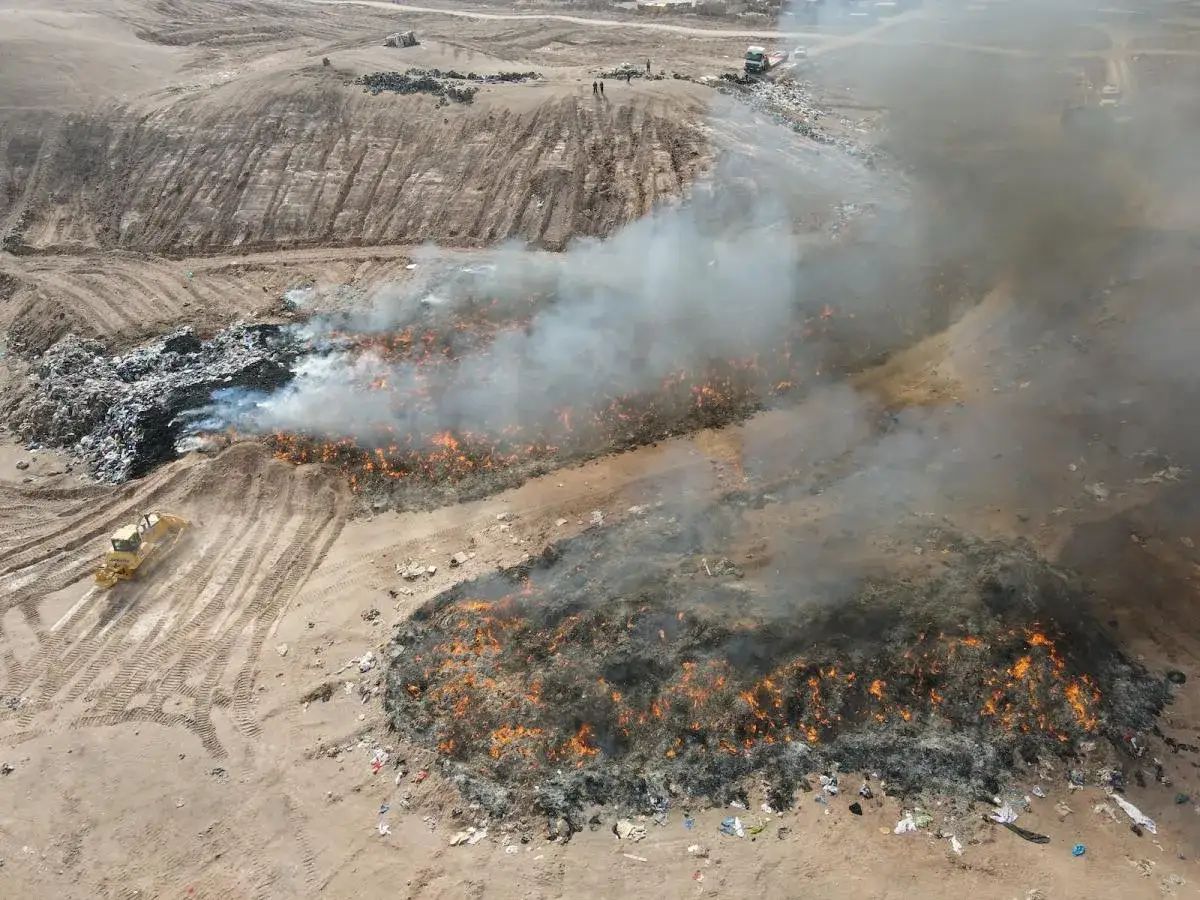 Vasta discarica di vestiti in Cile in fiamme, con fumo denso e un bulldozer che lavora tra i rifiuti.