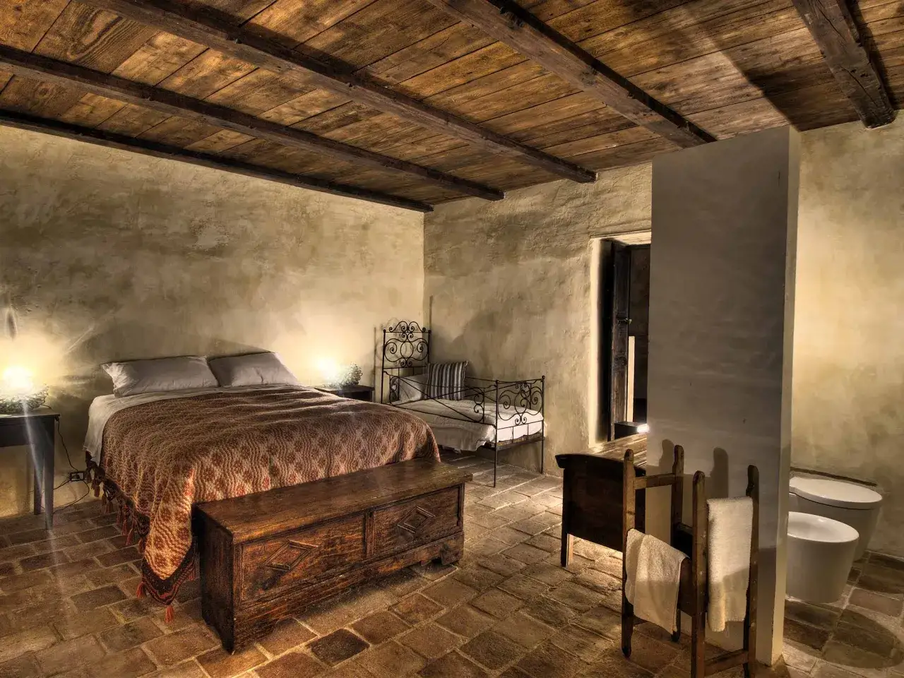Camera da letto rustica con soffitto in legno, letto matrimoniale, cassapanca antica e bagno. Un esempio di albergo diffuso significato.