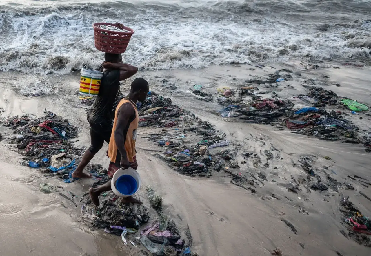 Spiaggia inquinata da rifiuti tessili, simbolo dei pro e contro del fast fashion. Due uomini camminano tra i detriti.