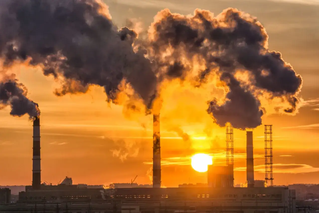 Fumi scuri escono da ciminiere industriali contro un tramonto arancione, simbolo di quali materie prime non sono rinnovabili.