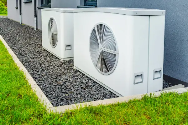 Due unità esterne di pompe di calore alta temperatura, bianche e moderne, installate su ghiaia accanto a un prato verde e a un edificio grigio.