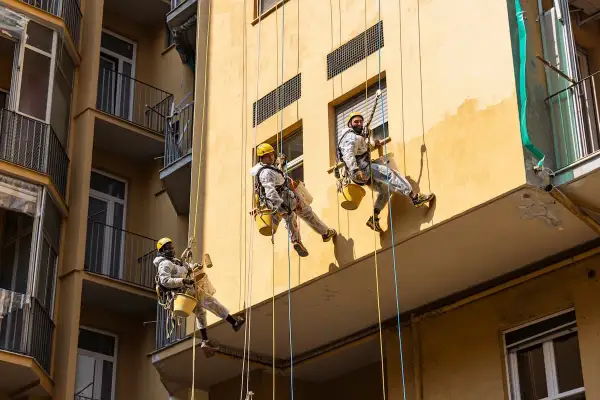 Lavoratori in corda applicano intonaco esterno tipi su un edificio giallo.
