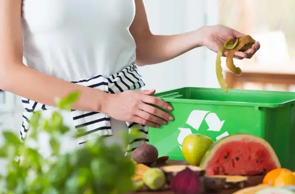 Mani che gettano bucce di frutta in un contenitore verde per ridurre i rifiuti. Frutta e verdura sul tavolo.